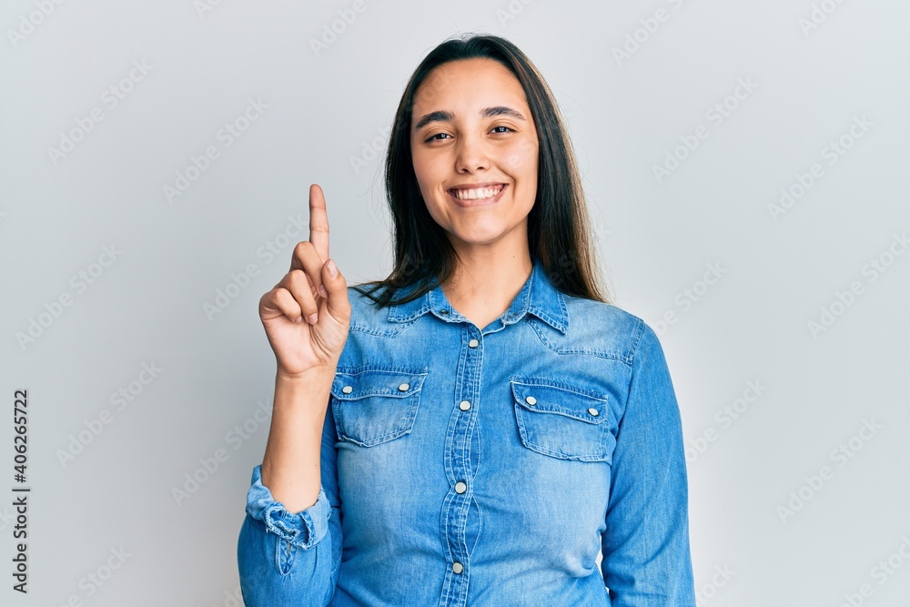 Young hispanic woman wearing casual denim jacket showing and pointing up with finger number one while smiling confident and happy. Young hispanic woman wearing casual denim jacket showing and pointing up with finger number one while smiling confident and happy.