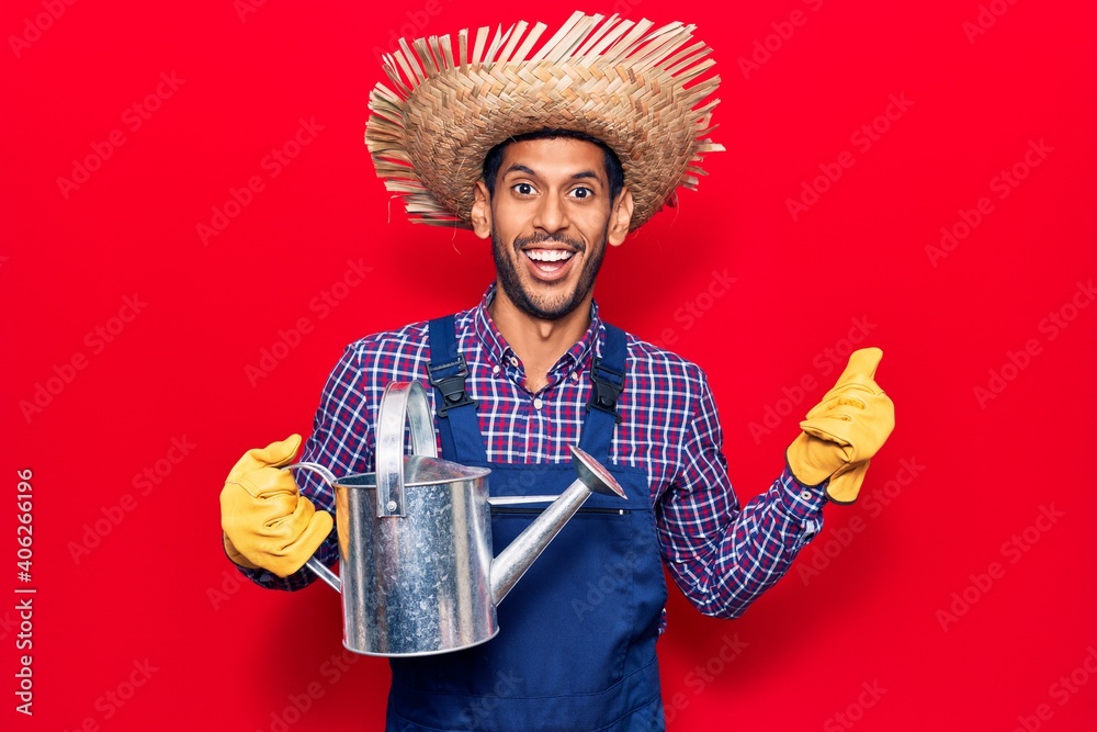 Young latin man wearing farmer hat and gloves holding watering can pointing thumb up to the side smiling happy with open mouth - obrazy, fototapety, plakaty 
