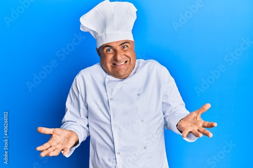 Mature middle east man wearing professional cook uniform and hat clueless and confused expression with arms and hands raised. doubt concept.