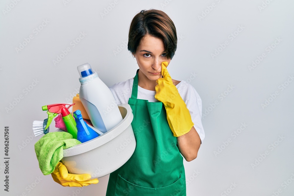 Young brunette woman with short hair wearing apron holding cleaning products pointing to the eye watching you gesture, suspicious expression