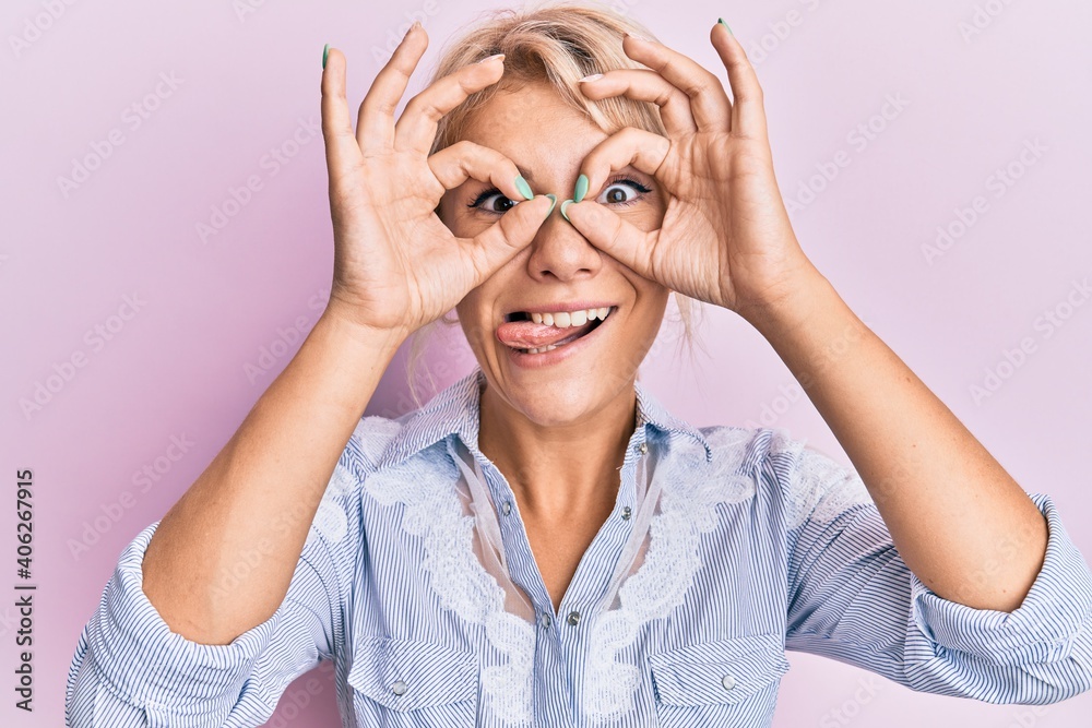 Young blonde girl wearing casual clothes doing ok gesture like binoculars sticking tongue out, eyes looking through fingers. crazy expression. Young blonde girl wearing casual clothes doing ok gesture like binoculars sticking tongue out, eyes looking through fingers. crazy expression.
