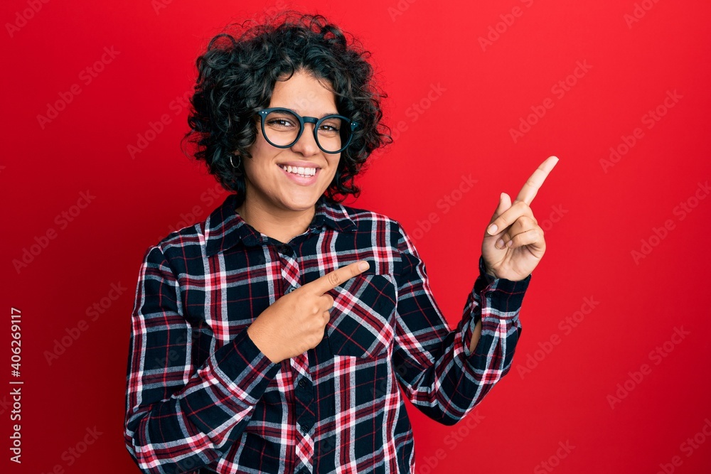 Young hispanic woman with curly hair wearing casual clothes and glasses smiling and looking at the camera pointing with two hands and fingers to the side. Young hispanic woman with curly hair wearing casual clothes and glasses smiling and looking at the camera pointing with two hands and fingers to the side.