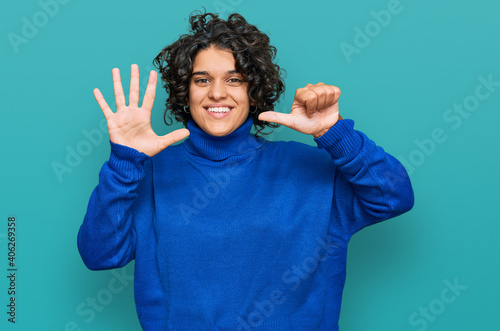 Young hispanic woman with curly hair wearing turtleneck sweater showing and pointing up with fingers number six while smiling confident and happy.