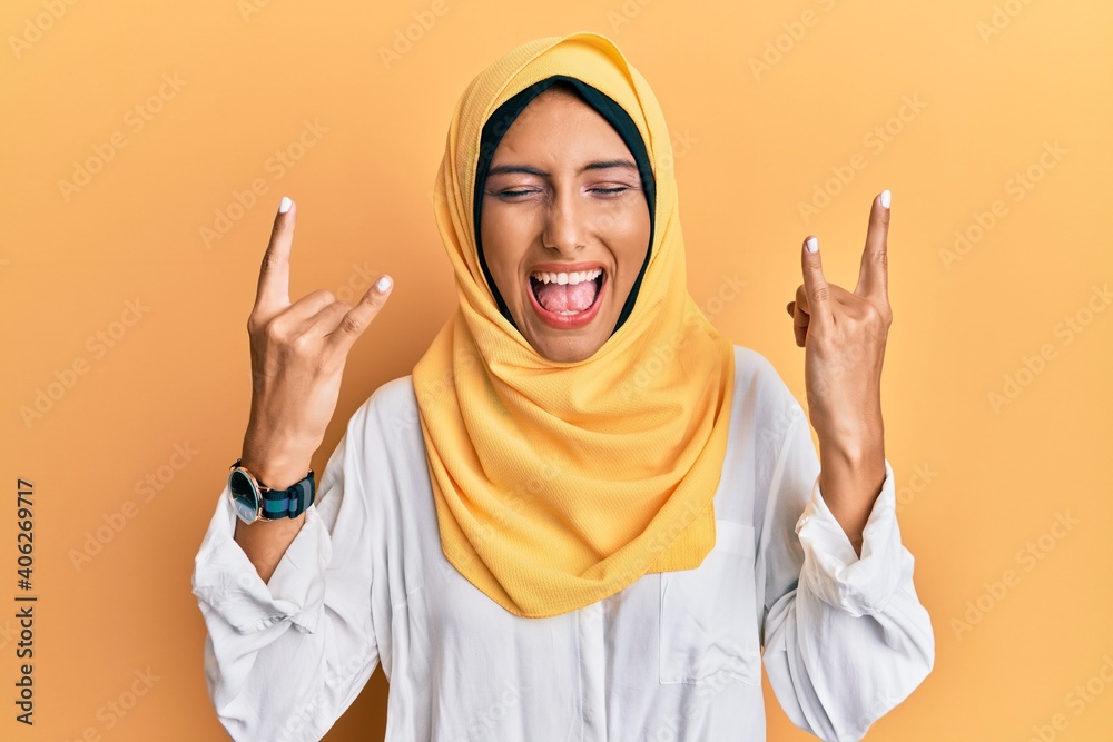 Young brunette arab woman wearing traditional islamic hijab scarf shouting with crazy expression doing rock symbol with hands up. music star. heavy music concept.