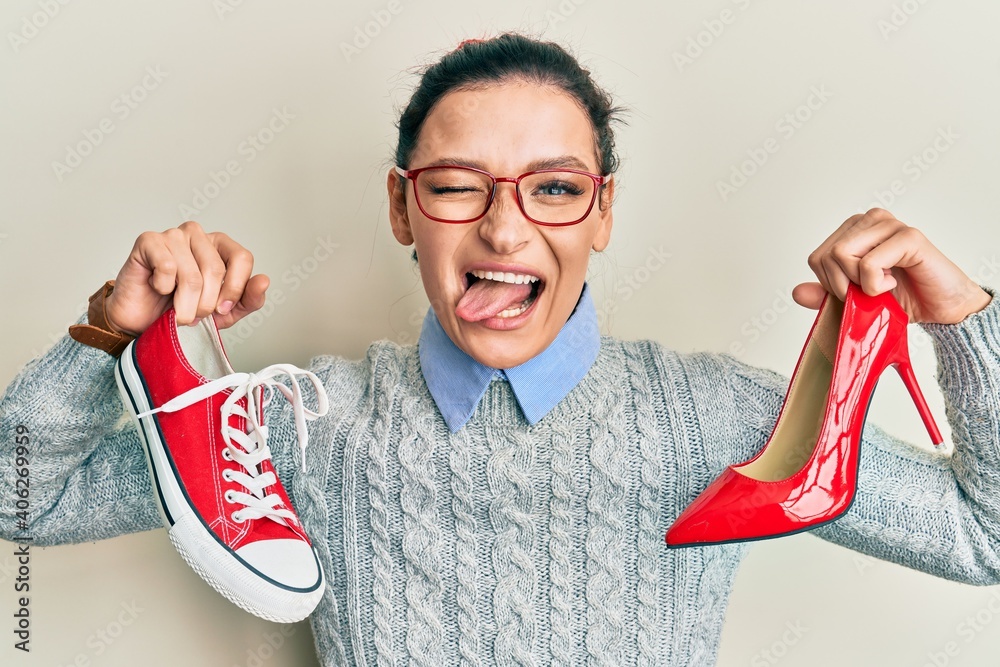 Young caucasian woman choosing high heel shoes and sneakers sticking tongue out happy with funny expression. - obrazy, fototapety, plakaty 