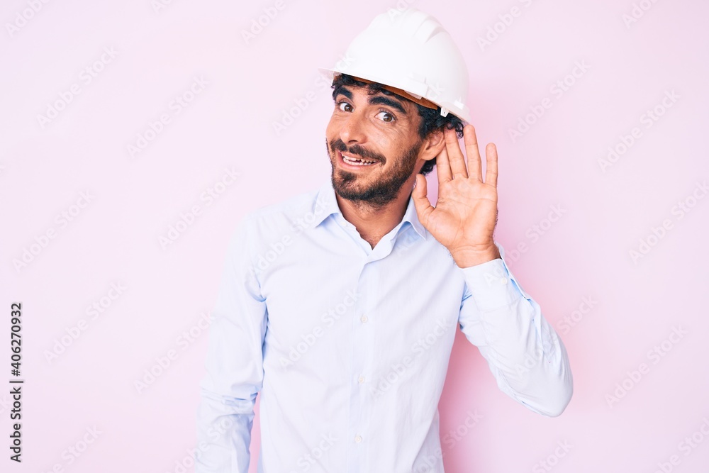 Handsome young man with curly hair and bear wearing architect hardhat smiling with hand over ear listening and hearing to rumor or gossip. deafness concept. Handsome young man with curly hair and bear wearing architect hardhat smiling with hand over ear listening and hearing to rumor or gossip. deafness concept.
