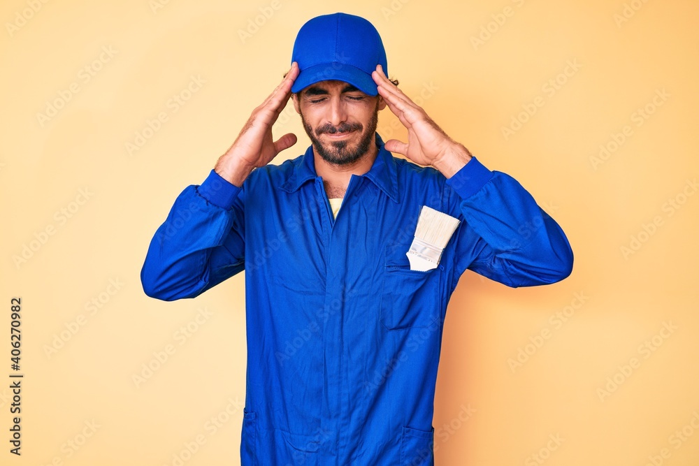 Handsome young man with curly hair and bear wearing builder jumpsuit uniform with hand on head for pain in head because stress. suffering migraine.