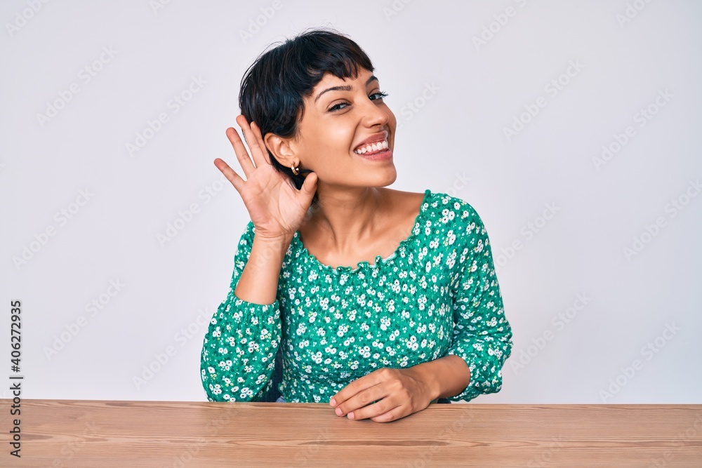 Beautiful brunettte woman wearing casual clothes sitting on the table smiling with hand over ear listening an hearing to rumor or gossip. deafness concept. Beautiful brunettte woman wearing casual clothes sitting on the table smiling with hand over ear listening an hearing to rumor or gossip. deafness concept.