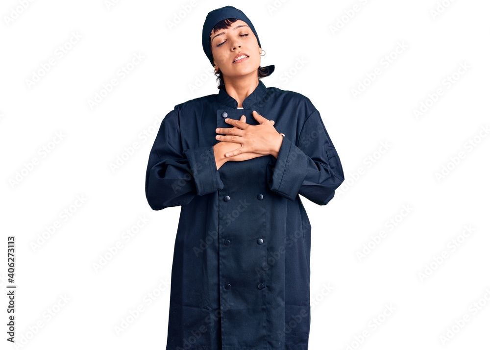 Young brunette woman with short hair wearing professional cook uniform smiling with hands on chest with closed eyes and grateful gesture on face. health concept. Young brunette woman with short hair wearing professional cook uniform smiling with hands on chest with closed eyes and grateful gesture on face. health concept.