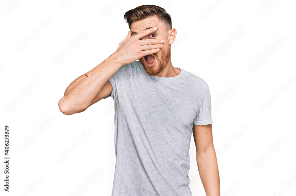 Young redhead man wearing casual grey t shirt peeking in shock covering face and eyes with hand, looking through fingers with embarrassed expression.