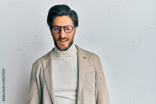 Young hispanic man wearing business jacket and glasses winking looking at the camera with sexy expression, cheerful and happy face.