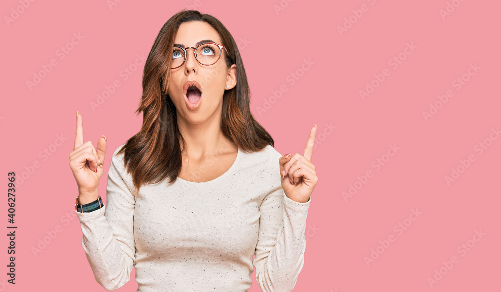 Young brunette woman wearing casual clothes and glasses amazed and surprised looking up and pointing with fingers and raised arms. Young brunette woman wearing casual clothes and glasses amazed and surprised looking up and pointing with fingers and raised arms.