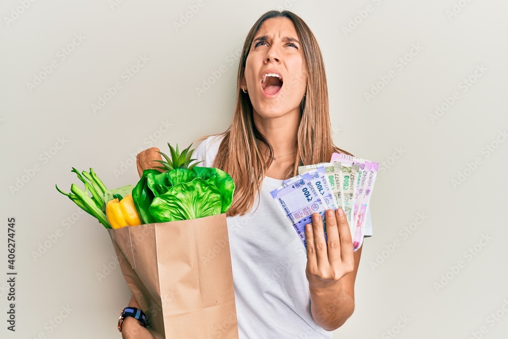 Brunette young woman holding groceries and indian rupee banknotes angry and mad screaming frustrated and furious, shouting with anger looking up. Brunette young woman holding groceries and indian rupee banknotes angry and mad screaming frustrated and furious, shouting with anger looking up.