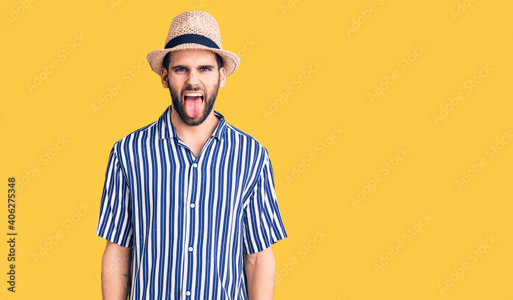 Young handsome man with beard wearing summer hat and striped shirt sticking tongue out happy with funny expression. emotion concept.