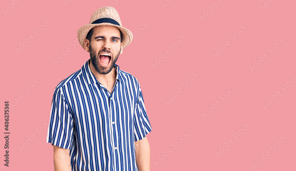 Young handsome man with beard wearing summer hat and striped shirt winking looking at the camera with sexy expression, cheerful and happy face. Young handsome man with beard wearing summer hat and striped shirt winking looking at the camera with sexy expression, cheerful and happy face.