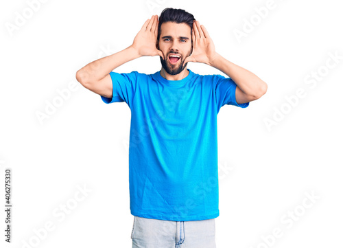 Young handsome man with beard wearing casual t-shirt smiling cheerful playing peek a boo with hands showing face. surprised and exited