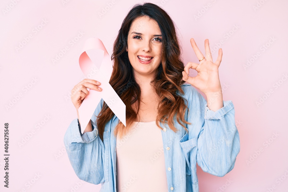 Young beautiful brunette woman holding pink cancer ribbon doing ok sign with fingers, smiling friendly gesturing excellent symbol Young beautiful brunette woman holding pink cancer ribbon doing ok sign with fingers, smiling friendly gesturing excellent symbol