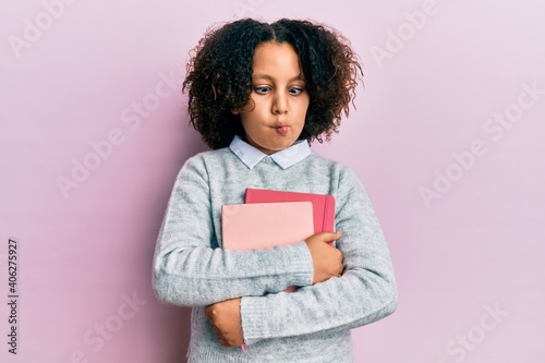 Young little girl with afro hair holding books making fish face with mouth and squinting eyes, crazy and comical.