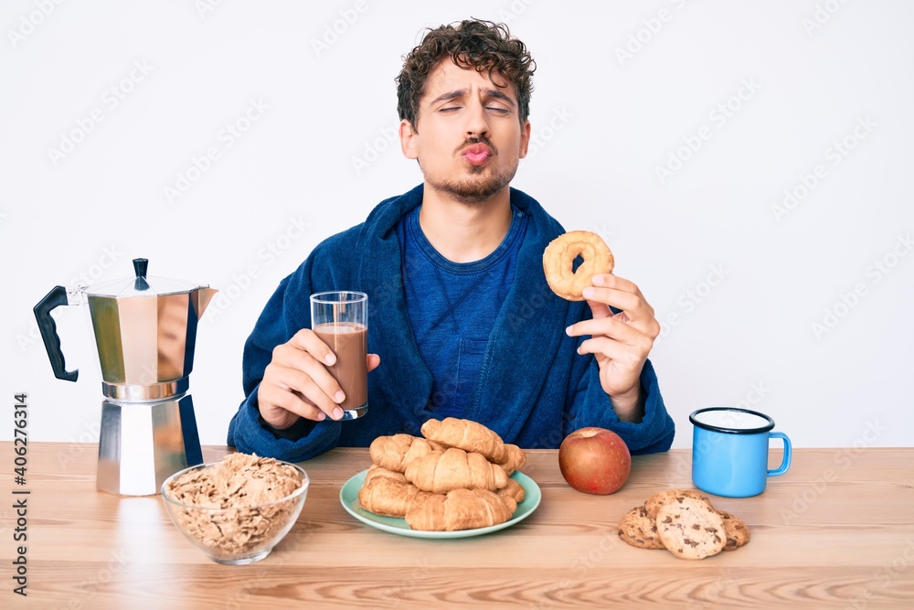 Young caucasian man with curly hair eating breakfast holding chocolate beverage and donut looking at the camera blowing a kiss being lovely and sexy. love expression. Young caucasian man with curly hair eating breakfast holding chocolate beverage and donut looking at the camera blowing a kiss being lovely and sexy. love expression.