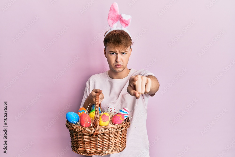 Young caucasian man wearing cute easter bunny ears holding wicker basket with colored eggs pointing with finger to the camera and to you, confident gesture looking serious