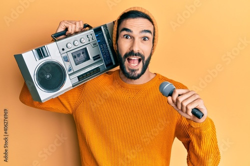 Young hispanic man holding boombox, listening to music singing with microphone celebrating crazy and amazed for success with open eyes screaming excited.