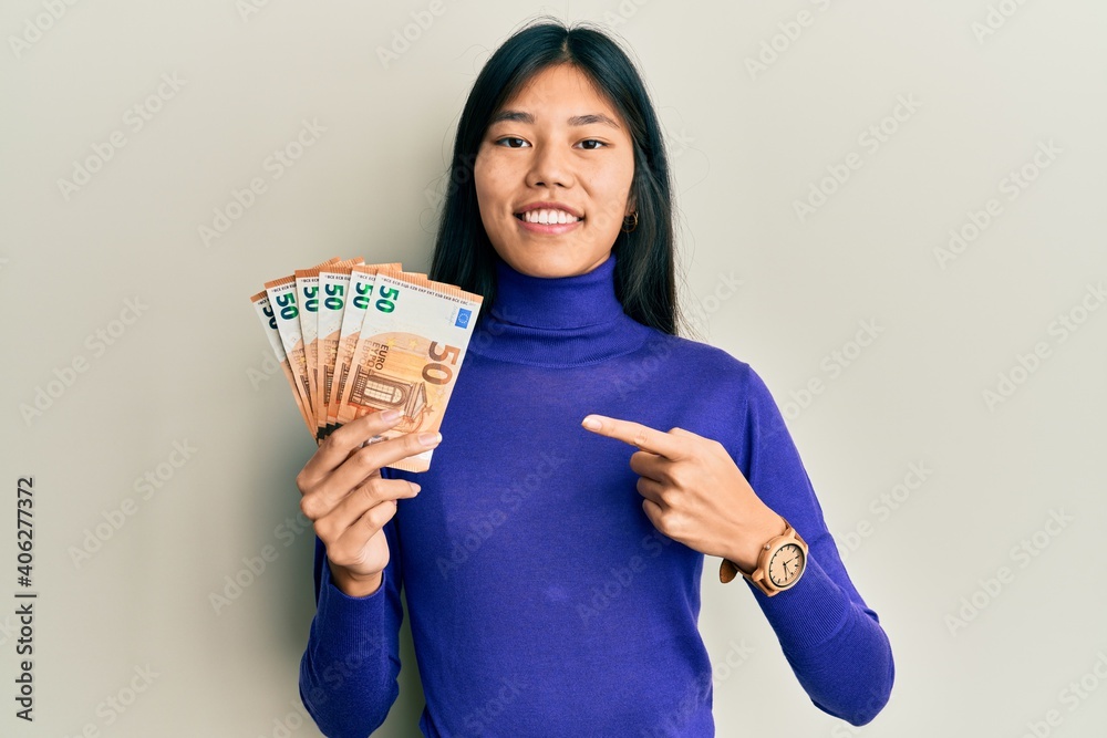 Young asian woman holding bunch of 50 euro banknotes smiling happy pointing with hand and finger Young asian woman holding bunch of 50 euro banknotes smiling happy pointing with hand and finger