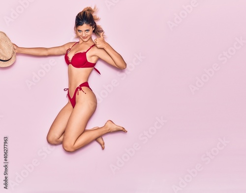Young beautiful girl on vacation wearing bikini smiling happy. Jumping with smile on face holding summer hat doing ok sign with thumb up over isolated pink background