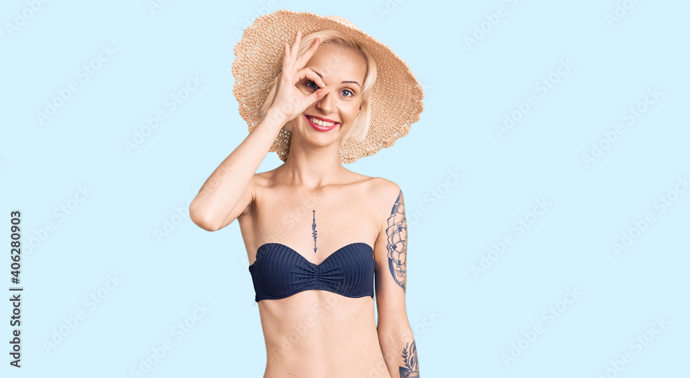 Young blonde woman with tattoo wearing bikini and summer hat doing ok gesture with hand smiling, eye looking through fingers with happy face.