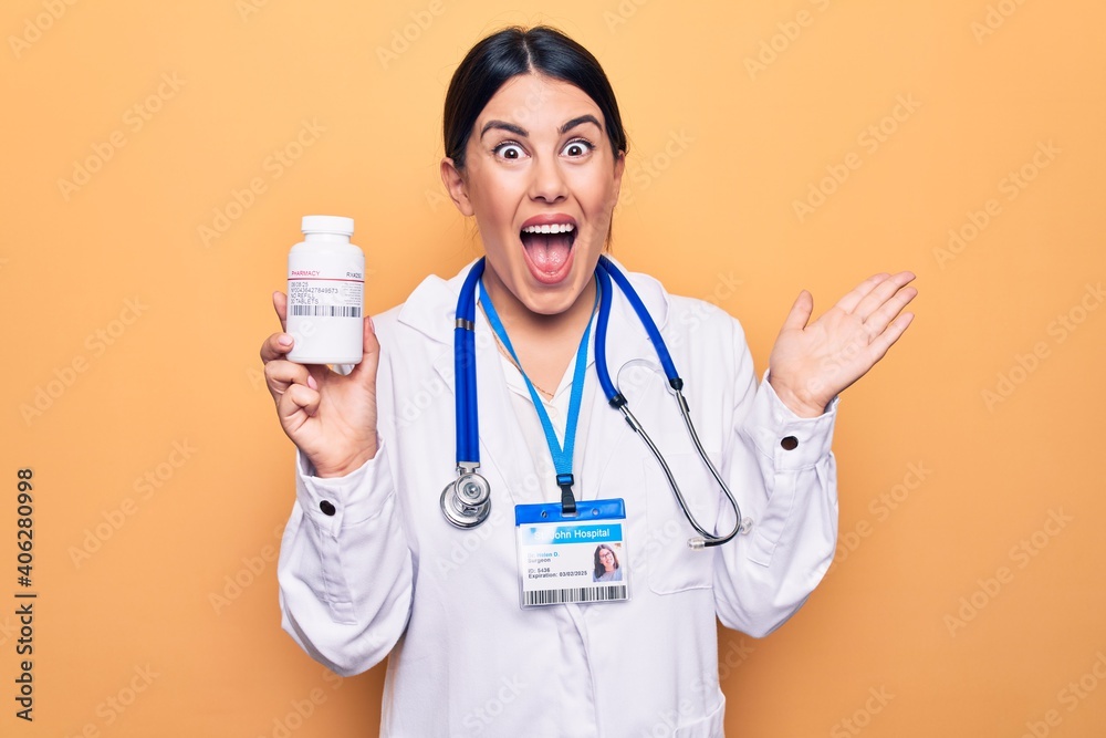 Young doctor woman wearing stethoscope holding bottle of pills over yellow background celebrating achievement with happy smile and winner expression with raised hand