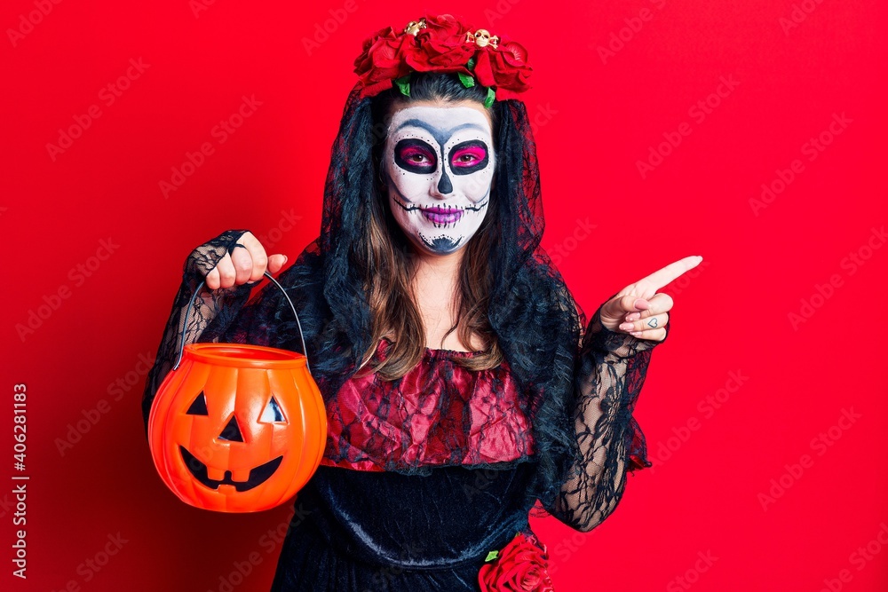 Young woman wearing day of the dead costume holding pumpkin smiling happy pointing with hand and finger to the side