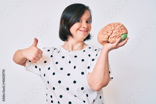 Brunette woman with down syndrome holding brain smiling happy and positive, thumb up doing excellent and approval sign