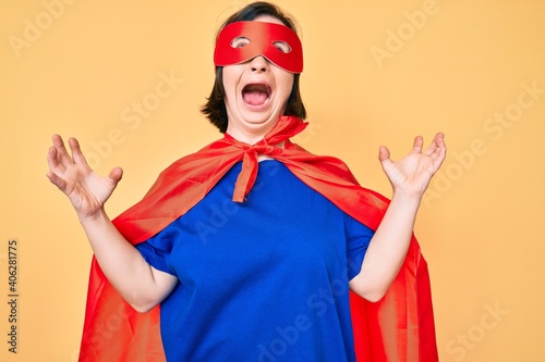 Brunette woman with down syndrome wearing super hero costume crazy and mad shouting and yelling with aggressive expression and arms raised. frustration concept.