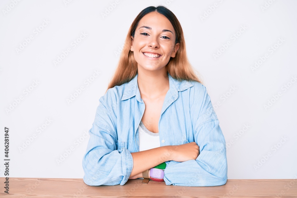 Young brunette woman sitting on the table wearing casual clothes happy face smiling with crossed arms looking at the camera. positive person. Young brunette woman sitting on the table wearing casual clothes happy face smiling with crossed arms looking at the camera. positive person.