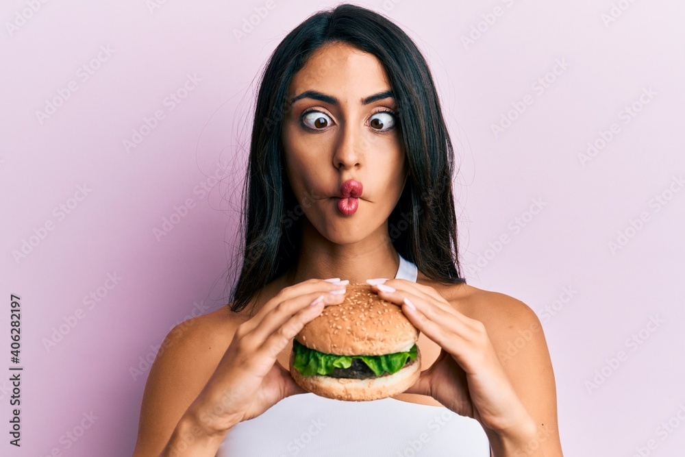 Beautiful hispanic woman eating a tasty classic burger making fish face with mouth and squinting eyes, crazy and comical.