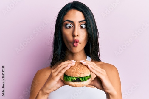 Beautiful hispanic woman eating a tasty classic burger making fish face with mouth and squinting eyes  crazy and comical.