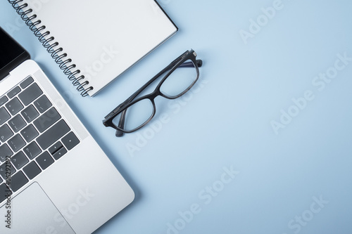 Blank notebook and pen on laptop computer with eye glasses on blue background