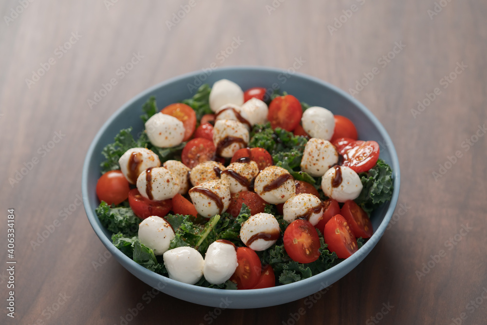 Blue bowl of salad with mozzarella, cherry tomatoes and kale, seasoned with balsamic sauce on walnut wood table Blue bowl of salad with mozzarella, cherry tomatoes and kale, seasoned with balsamic sauce on walnut wood table