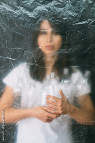 Plastic pollution. Defocused silhouette. Garbage collection. Zero waste. Volunteer woman holding used bottles behind wrinkled texture polyethylene film isolated on dark background out of focus.