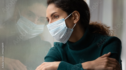 Upset young Caucasian woman in medical facial mask look in window feel lonely distressed sick with covid-19. Unhappy female in facemask suffer from coronavirus. Quarantine, corona pandemic concept. photo