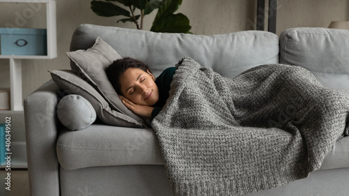 Happy millennial Caucasian woman lying under blanket on cozy couch at home sleeping or dreaming. Smiling calm young female relax rest on sofa in living room, relieve negative emotions daydreaming. photo
