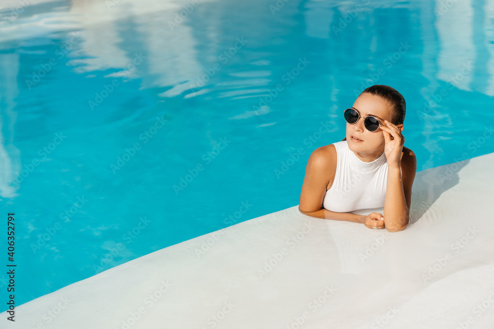 Young woman relaxing in swimming pool on summer vacation. Blonde caucasian model in white bikini floating in water.