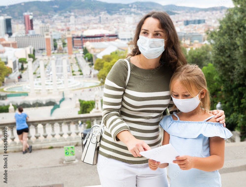Young woman in medical mask pointing to sightseeing with daughter at the street among architecture at Barcelona