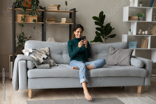 Smiling young Caucasian woman sit relax on couch in living room talk on video call. Happy millennial female rest on sofa at home have webcam digital virtual communication on cellphone.