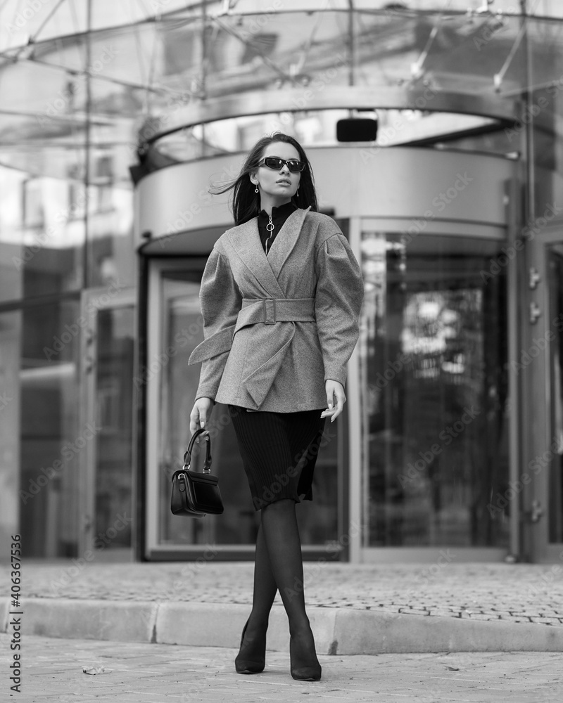Pretty tall caucsian woman with long straight brunette hair in gray short coat, black skirt, tights, sunglasses and shoes walking city street near modern building
