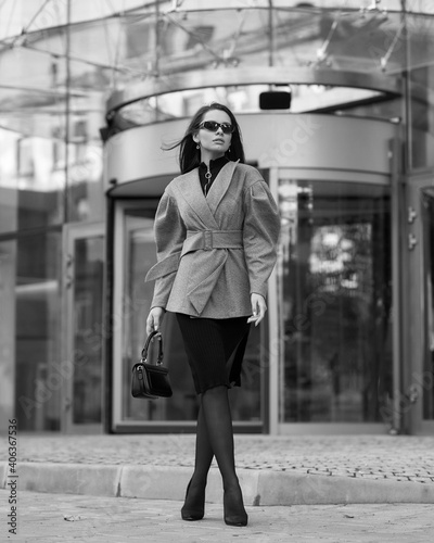 Pretty tall caucsian woman with long straight brunette hair in gray short coat, black skirt, tights, sunglasses and shoes walking city street near modern building photo