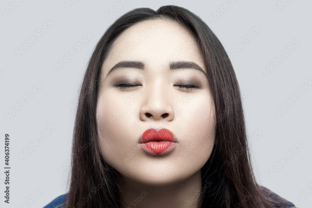 Portrait of in love beautiful brunette asian young woman in casual blue denim jacket with makeup standing with closed eyes and sending kiss. indoor studio shot, isolated on light grey background.