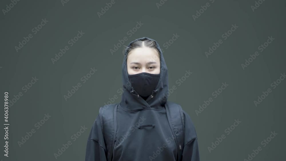 Angry rebel asian girl in black mask and hoodie protestor activist posing on gray studio background Angry rebel asian girl in black mask and hoodie protestor activist posing on gray studio background