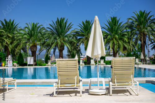 Two white plastic sun loungers and parasol stand near the pool with turquoise water on the pallm trees background © Oksana