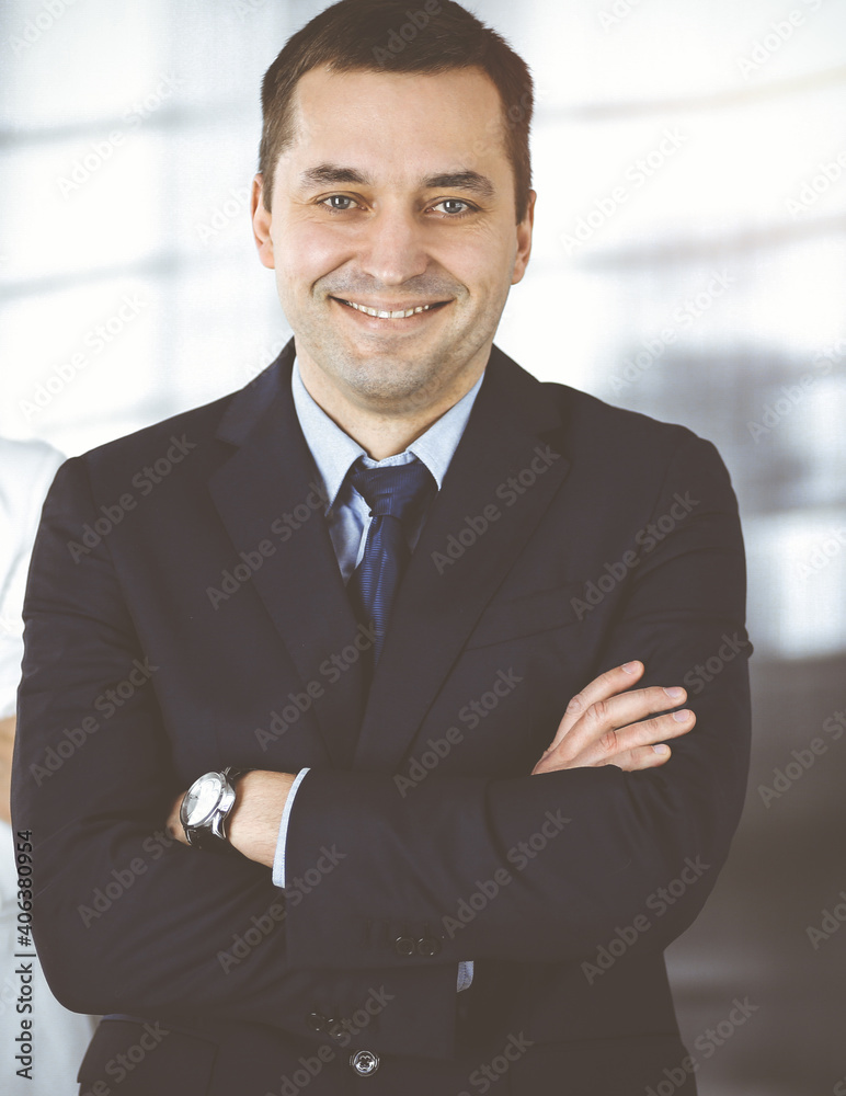 Portrait of a friendly middle aged businessman in a dark blue suit, standing with crossed arms together with a colleague in a sunny modern office. Business concept Portrait of a friendly middle aged businessman in a dark blue suit, standing with crossed arms together with a colleague in a sunny modern office. Business concept