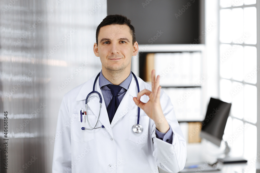 Friendly male doctor standing straight with OK sign in clinic near his working place, closeup. Perfect medical service in hospital. Medicine and healthcare concept Friendly male doctor standing straight with OK sign in clinic near his working place, closeup. Perfect medical service in hospital. Medicine and healthcare concept